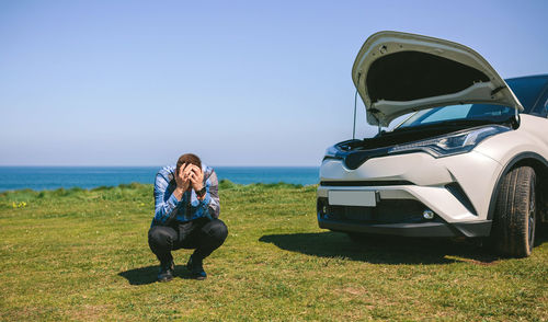 Frustrated man crouching against broken down car at beach