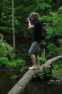 Side view full length of photographer photographing while standing on fallen tree at forest