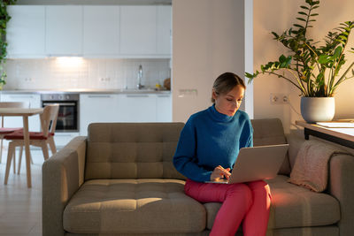 Young freelancer sitting on cozy sofa works on laptop and makes business project in apartment