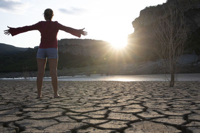 Backlit woman at the edge of a lake with her arms outstretched.