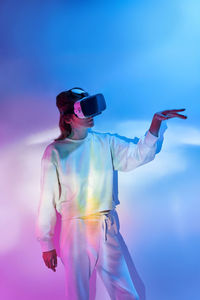 Woman in modern vr goggles looking at raised arm while exploring virtual reality near wall in room with glowing light