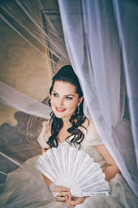 Close-up of smiling bride holding hand fan while sitting on bed at home