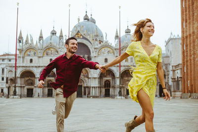 Happy couple running while holding hands in city against buildings and sky