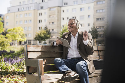 Senior man with tablet pc enjoying music listening through wireless headphones on bench