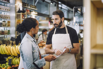 Mature salesman discussing over document with female customer at delicatessen