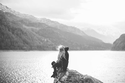 Young woman sitting on rock by lake against sky