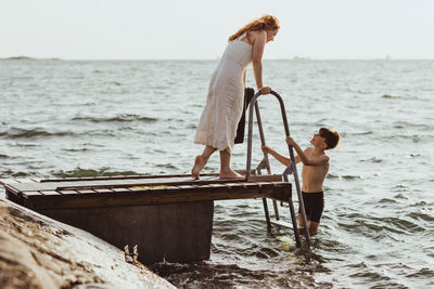 Side view of woman talking while son standing on ladder in sea