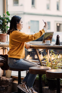 Full length side view of female blogger taking selfie sitting at cafe