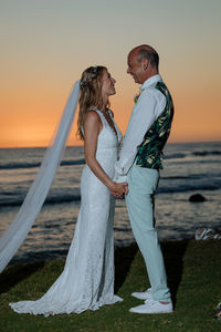 Bride and groom standing by sea during sunset