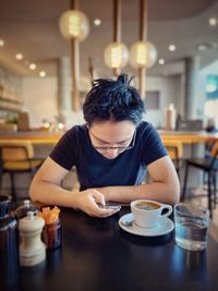 Young asian man in eyeglasses using mobile phone while drinking coffee at table in cafe.