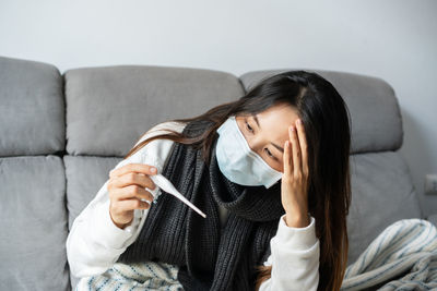 Woman wearing protective face mask examining thermometer at home