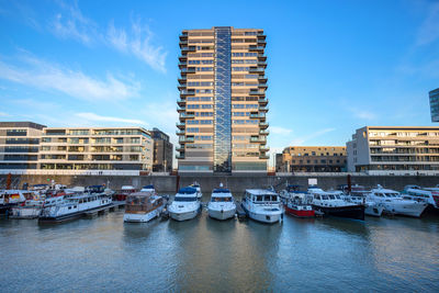 Boats moored at harbor by buildings against blue sky