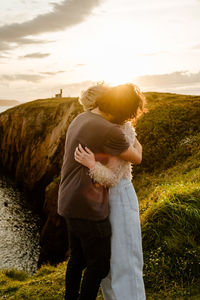 Side view of unrecognizable boyfriend and girlfriend embracing each other while standing on grassy cliff against cloudy sundown sky in aviles, spain