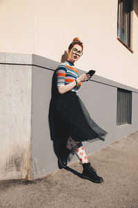 Full length of young woman using mobile phone while standing by wall