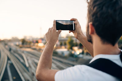 Young man photographing through smart phone over railroad tracks in city