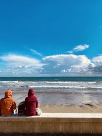 Rear view of people seating in jackets at beach against the water and sky vertical
