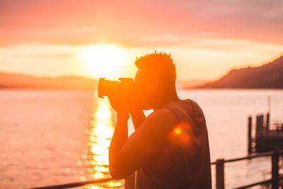 Side view of man photographing with digital camera by lake against orange sky during sunset