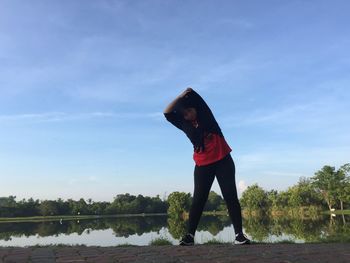 Full length of woman exercising while bending against lake and sky