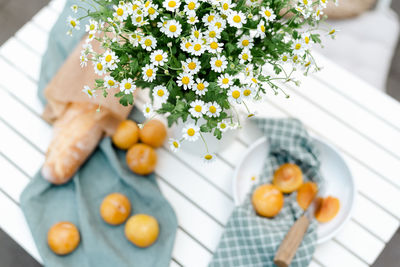 Bouquet of chamomile in a white jug, ripe yellow plums, and fresh baguette on a white table.
