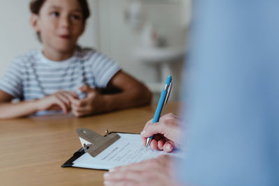 Cropped hands of doctor writing prescription while boy sitting at desk in clinic