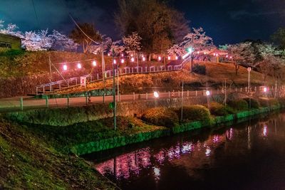 Illuminated street by lake against sky at night