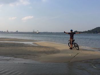 Full length of man with arms outstretched standing by bicycle at beach against sky