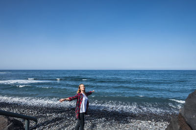 Woman with arms outstretched standing at beach against clear blue sky