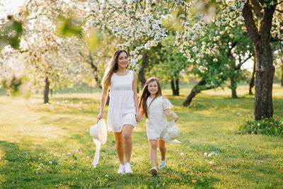 Mom and her daughter in white dresses run through a blooming apple orchard in spring on a sunny day