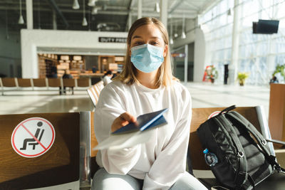 Young woman traveler in medical mask sits in the waiting room and holds out passport and ticket