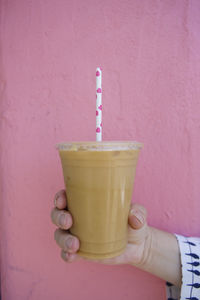View of a hand holding an iced latte  against a pink wall with a paper straw with pink hearts