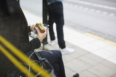 Midsection of businesswoman eating sandwich while sitting with smart phone at bus stop in city