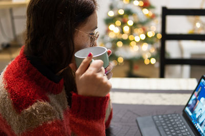 Woman making video call taking a cup of tea with illuminated christmas tree in the background