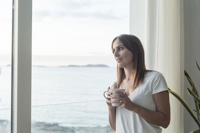 Woman having coffee while looking through window at home