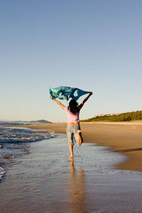 Rear view of mid adult woman holding scarf while walking at beach against clear sky