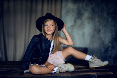 Fashionable girl model in a leather jacket skirt and shoes sit against the wall in the studio