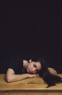 Portrait of young woman relaxing on wooden bench against black wall at home