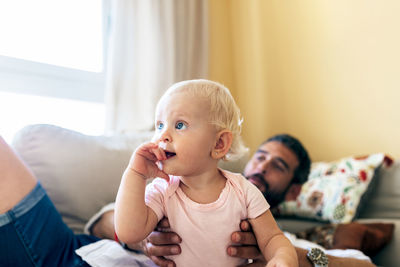 Happy bearded father holding cute laughing baby while resting on couch at home