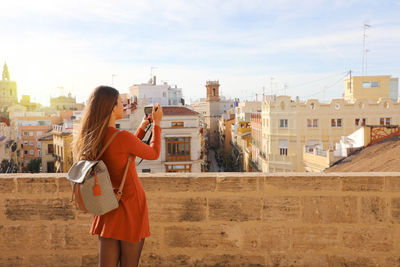 Woman photographing with camera while standing in city against sky