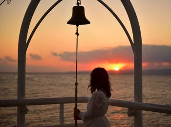 Rear view of woman standing by railing against sea during sunset