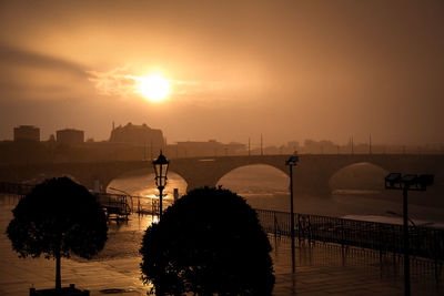 Silhouette bridge over river in city against sky during sunset