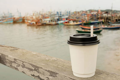 White hot coffee paper cup with plastic black lid on wooden bar with nature sea view as background