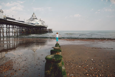 Rear view of woman standing on moss covered wooden post at beach by eastbourne pier against sky