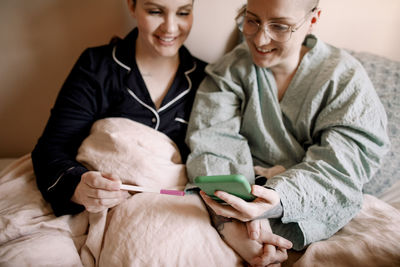 Happy lesbian couple checking pregnancy test results with smart phone while sitting on bed at home