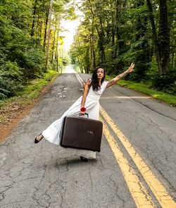Full length of young woman with suitcase gesturing while standing on road