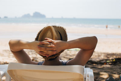 Midsection of mid adult man with hands behind head relaxing on deck chair at beach against sky