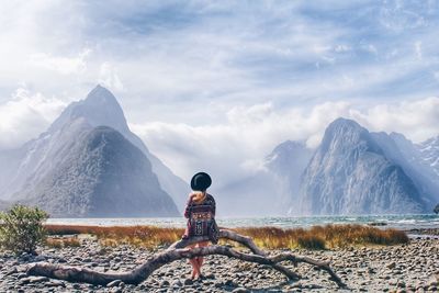 Young woman sitting on driftwood at lakeshore against snowcapped mountains