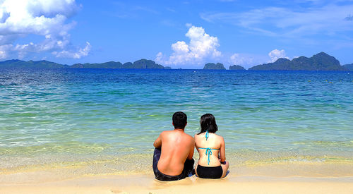 Rear view of couple sitting at beach against cloudy sky
