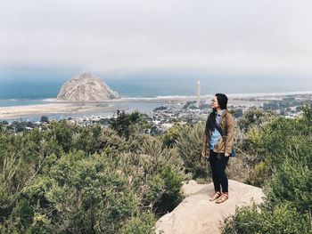 Young woman standing on rock amidst trees by sea against sky