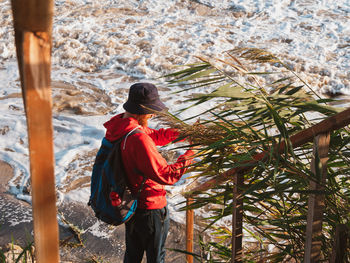 Bearded man in red hoody with backpack standing against sea with reeds. millennial guy travel alone.