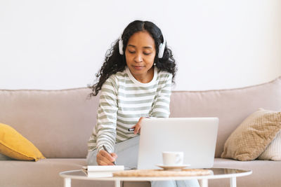 African-american female taking notes in notebook while listening to online course in earphones